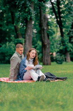 Parkta, kalın elbiseli bir adam ve kız, mavi bir battaniyeyle kaplı, kareli bir battaniyenin üzerinde kitap okuyan bir kıza sarılıyorlar.