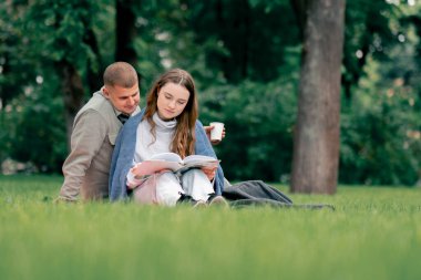Parkta, kalın elbiseli bir adam ve kız, mavi bir battaniyeyle kaplı, kareli bir battaniyenin üzerinde kitap okuyan bir kıza sarılıyorlar.