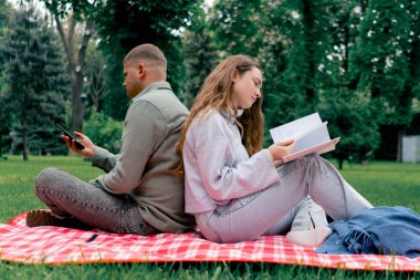 Parkta kalın elbiseli bir adam ve kız, kareli bir battaniyenin üzerinde dinleniyorlardı. Kız kitap okuyordu ve adam telefonda sırt sırta konuşuyordu.