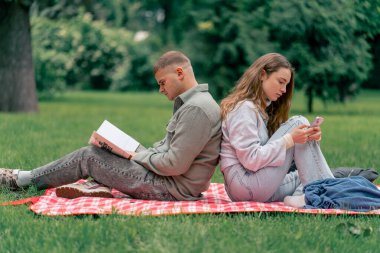 Parktaki adam, üzerinde kalın giysiler olan bir kızla kareli bir battaniyede kitap okuyor ve kız telefonda, arka arkaya modern gençlerle konuşuyor.