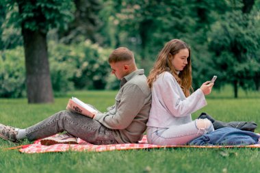Parktaki adam, üzerinde kalın giysiler olan bir kızla kareli bir battaniyede kitap okuyor ve kız telefonda, arka arkaya modern gençlerle konuşuyor.