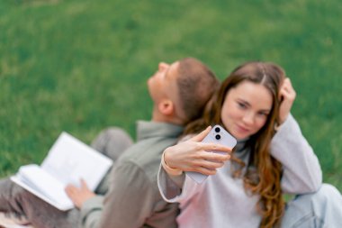 Parktaki adamın yanında kalın elbiseli bir kız ve kareli bir battaniyeyle adam kitap okuyor ve kız arka arkaya telefonda modern gençlerin fotoğraflarını çekiyor.