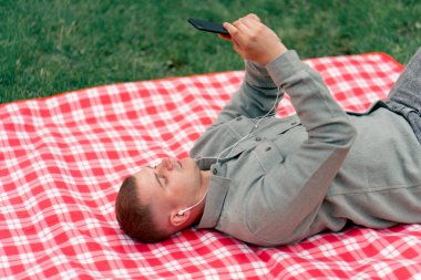 Parkta kırmızı kareli battaniyeli bir adam telefonundan beyaz kulaklıklarla müzik dinliyor.