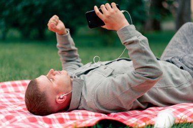 Parkta kırmızı kareli battaniyeli bir adam telefonundan beyaz kulaklıklarla müzik dinliyor.