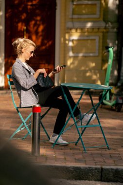 Şehirde güneşli bir caddede yol kenarında bir kahve dükkanında demir bir masa ve nane rengi sandalyeler var.