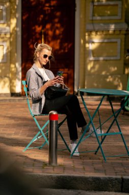 Şehirde güneşli bir caddede yol kenarında bir kahve dükkanında demir bir masa ve nane rengi sandalyeler var.