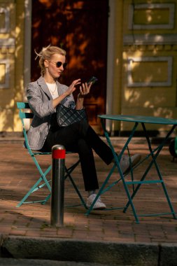 Şehirde güneşli bir caddede yol kenarında bir kahve dükkanında demir bir masa ve nane rengi sandalyeler var.