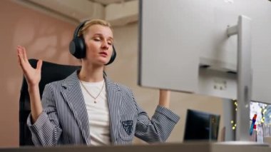 Parlak bir ofiste sarışın genç bir çalışan beyaz ekranda çalışıyor. Masanın üzerinde modern ofis ışınları çalışıyor. Büyük siyah kulaklıklar üzerinde çalışıyor.