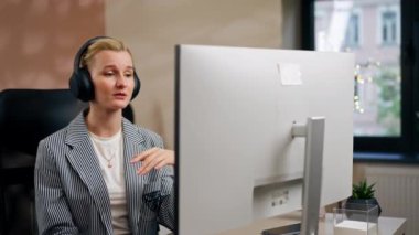 Parlak bir ofiste sarışın genç bir çalışan beyaz ekranda çalışıyor. Masanın üzerinde modern ofis ışınları çalışıyor. Büyük siyah kulaklıklar üzerinde çalışıyor.