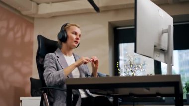 Parlak bir ofiste sarışın genç bir çalışan beyaz ekranda çalışıyor. Masanın üzerinde modern ofis ışınları çalışıyor. Büyük siyah kulaklıklar üzerinde çalışıyor.