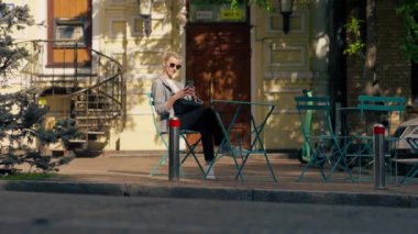 Şehirde güneşli bir caddede yol kenarında bir kahve dükkanında demir bir masa ve nane rengi sandalyeler var.