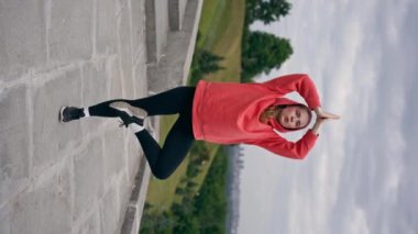 Parktaki dikey videoda turuncu kapüşonlu genç bir kız meditasyon yapıyor. Hava bulutlu.