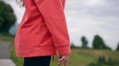 Parkta turuncu kapüşonlu genç bir kız, meditasyon yaparak ısınıyor. Hava bulutlu.