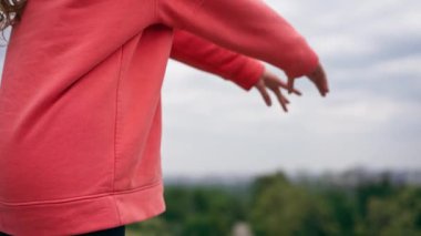 Parkta turuncu kapüşonlu genç bir kız, meditasyon yaparak ısınıyor. Hava bulutlu.