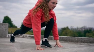 Parkta turuncu kapüşonlu genç bir kız, meditasyon yaparak ısınıyor. Hava bulutlu.