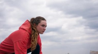 Parkta turuncu kapüşonlu genç bir kız, meditasyon yaparak ısınıyor. Hava bulutlu.