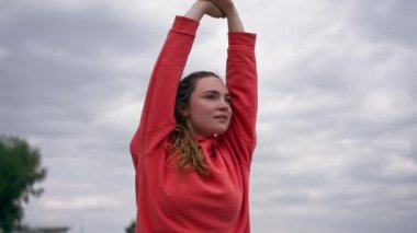 Parkta turuncu kapüşonlu genç bir kız, meditasyon yaparak ısınıyor. Hava bulutlu.