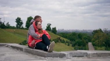 Parkta birbirine aşık genç bir çift spor yaptıktan sonra beton bir korkulukta sarılıp şehri seyrediyor, bulutlu bir hafta sonu