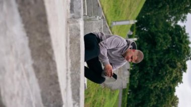 Parktaki dikey videoda gri kazaklı genç bir adam taş bir merdivende oturmuş telefon bulutlu havada sohbet ediyor.