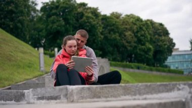 Parkta birbirine aşık genç bir çift spor yaptıktan sonra beton bir korkulukta sarılıyorlar, tablet bulutlu bir havada haberleri izliyorlar ortak bir hafta sonu.