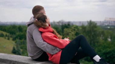 Parkta birbirine aşık genç bir çift spor yaptıktan sonra beton bir korkulukta sarılıp şehri seyrediyor, bulutlu bir hafta sonu