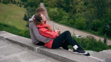 Parkta birbirine aşık genç bir çift spor yaptıktan sonra beton bir korkulukta sarılıp şehri seyrediyor, bulutlu bir hafta sonu