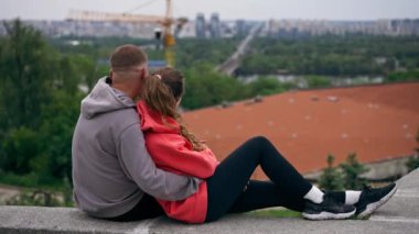 Parkta birbirine aşık genç bir çift spor yaptıktan sonra beton bir korkulukta sarılıp şehri seyrediyor, bulutlu bir hafta sonu