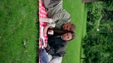 Dikey video. Parkta, kalın elbiseli bir kız ve erkek, mavi bir battaniyeye sarılmış, kitap okuyan bir kıza sarılıyorlar.