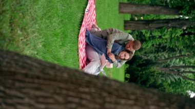 Dikey video. Parkta, kalın elbiseli bir kız ve erkek, mavi bir battaniyeye sarılmış, kitap okuyan bir kıza sarılıyorlar.