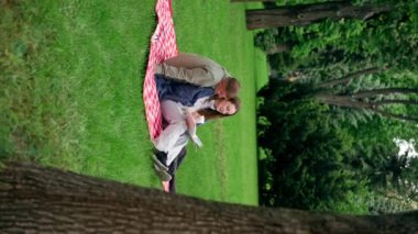 Dikey video. Parkta, kalın elbiseli bir kız ve erkek, mavi bir battaniyeye sarılmış, kitap okuyan bir kıza sarılıyorlar.