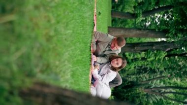 Dikey video. Parkta, kalın elbiseli bir kız ve erkek, mavi bir battaniyeye sarılmış, kitap okuyan bir kıza sarılıyorlar.