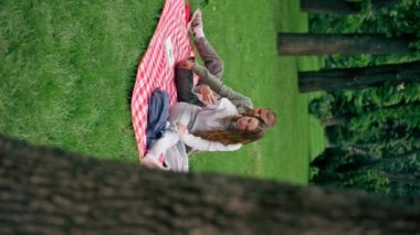 Dikey video. Parktaki adam, üzerinde kalın giysiler olan bir kızla kareli bir battaniyede kitap okuyor ve kız arka arkaya telefonla konuşuyor. Modern gençlik fotoğrafı yapıyor.