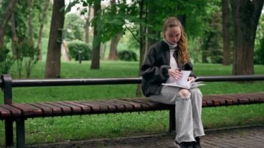 Parkta siyah ceketli, saçları birbirine dolanmış, yalnız bir kız bir bankta oturmuş kitap okuyor.