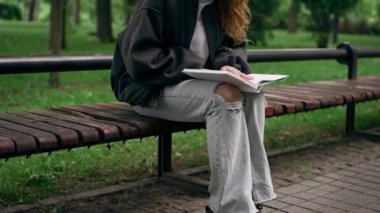 Parkta siyah ceketli, saçları birbirine dolanmış, yalnız bir kız bir bankta oturmuş kitap okuyor.