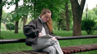 Parkta siyah ceketli, saçları birbirine dolanmış, yalnız bir kız bir bankta oturmuş kitap okuyor.
