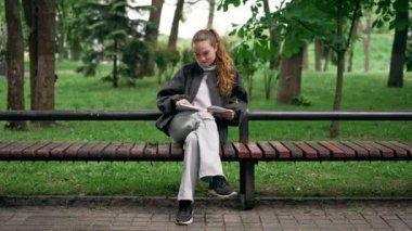 Parkta siyah ceketli, saçları birbirine dolanmış, yalnız bir kız bir bankta oturmuş kitap okuyor.