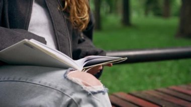 Parkta, siyah ceketli, saçları birbirine dolanmış, yalnız genç bir kız bir bankta oturmuş kitap okuyor.