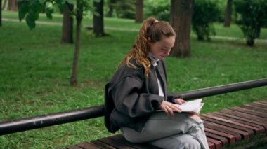 Parkta siyah ceketli, saçları birbirine dolanmış, yalnız bir kız bir bankta oturmuş kitap okuyor.