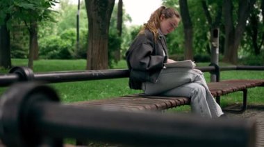 Parkta siyah ceketli, saçları birbirine dolanmış, yalnız bir kız bir bankta oturmuş kitap okuyor.