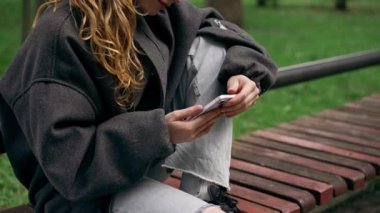 Parkta, siyah ceketli, saçları kabarık, cep telefonuyla mesajlaşan yalnız bir kız.