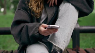 Parkta, siyah ceketli, saçları kabarık, cep telefonuyla mesajlaşan yalnız bir kız.