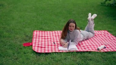 Parkta, kırmızı bir battaniyenin üzerinde bir kız başını dayayıp büyüleyici bir şekilde rahatlatıcı bir hafta sonu kitabı okuyor.