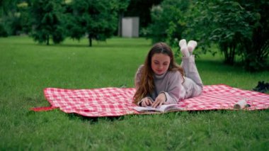 Parkta, kırmızı bir battaniyenin üzerinde bir kız başını dayayıp büyüleyici bir şekilde rahatlatıcı bir hafta sonu kitabı okuyor.