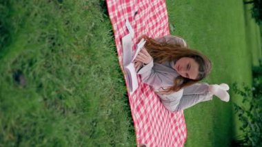 Parkta kırmızı bir battaniyeye sarılı dikey bir video. Bir kız başını kaldırıp kitap okuyor. Büyüleyici bir şekilde rahatlatıcı bir hafta sonu.