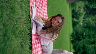 Parkta kırmızı bir battaniyeye sarılı dikey bir video. Bir kız başını kaldırıp kitap okuyor. Büyüleyici bir şekilde rahatlatıcı bir hafta sonu.