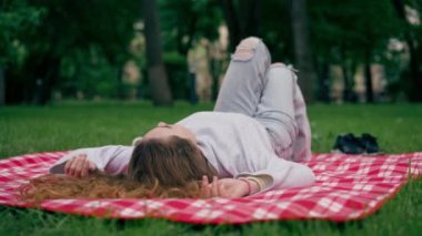 Parkta kırmızı kareli bir battaniyenin üzerinde elleri başının arkasında mavi gözlü bir kız yatıyor.