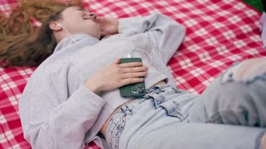 Parkta kırmızı kareli battaniyenin üzerinde mavi gözlü bir kız sırt üstü uzanmış saçlarını açmış müzik dinliyor.