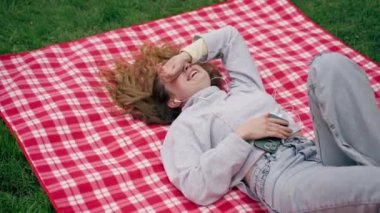 Parkta kırmızı kareli battaniyenin üzerinde mavi gözlü bir kız sırt üstü uzanmış saçlarını açmış müzik dinliyor.