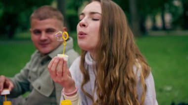 Doğadaki bir parkta, kırmızı kareli battaniyeye oturmuş sabun köpüklerini çocukluğa geri püskürten genç bir çift.