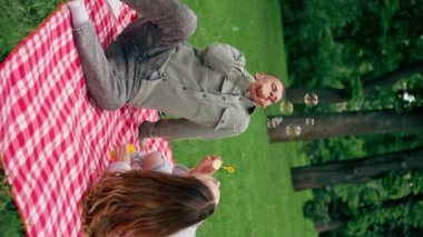 Doğadaki bir parkta kırmızı kareli battaniyeye oturmuş sabun köpüklerini çocukluğa geri püskürten genç bir çift.